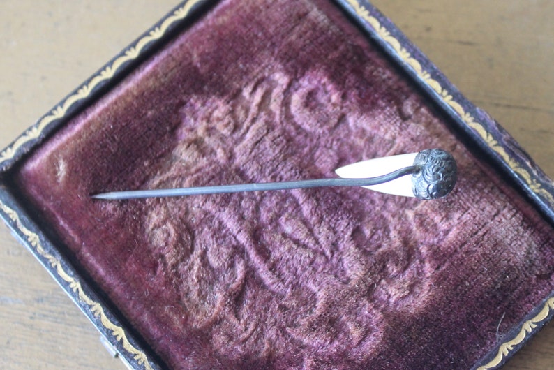 May include: A silver and white pin with a decorative floral design on the top. The pin is sitting in a velvet-lined box.