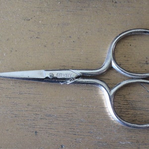 May include: Silver sewing scissors with a pointed tip and two circular handles. The scissors have "GERMANY" engraved on the blade.