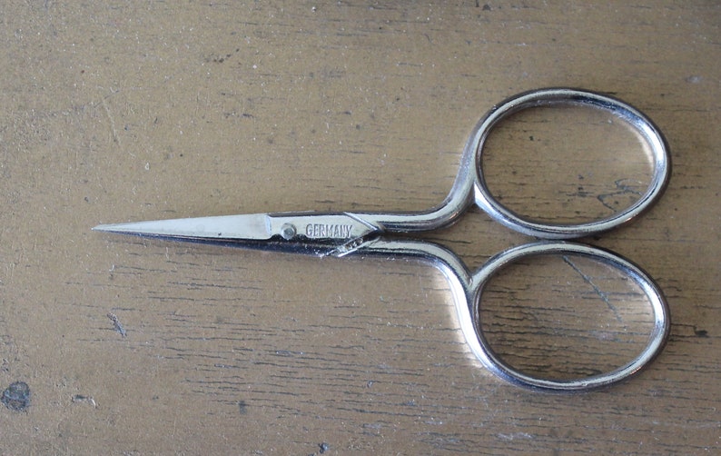 May include: Silver sewing scissors with a pointed tip and two circular handles. The scissors have "GERMANY" engraved on the blade.
