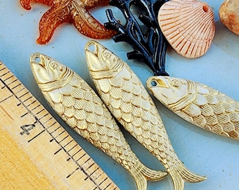 May include: Three gold-colored fish charms with detailed scales and a raised fin design. The charms are on a blue surface with a wooden ruler in the background.