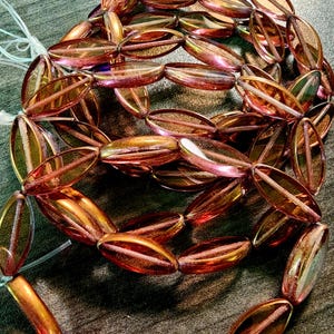 May include: A strand of translucent, elongated oval glass beads featuring a metallic, iridescent finish that shifts between shades of copper, bronze, and deep magenta. Each bead has a distinct central ridge, strung together on a thin, clear filament.