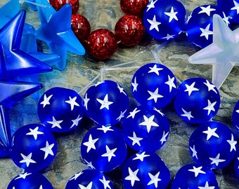May include: A collection of round beads featuring a blue base with white star patterns, interspersed with red glittery spherical beads and translucent blue star-shaped charms. The beads are arranged in a cluster, showcasing a mix of textures and colors.