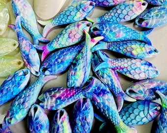 May include: A collection of colorful, fish-shaped beads in shades of blue, purple, pink, and green. The beads have a textured, scale-like pattern and are arranged in a scattered composition. Some beads are iridescent, reflecting light.