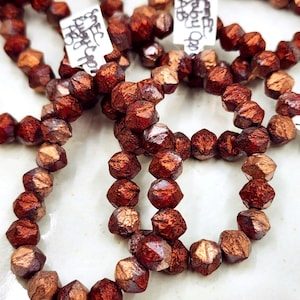 May include: A collection of faceted, metallic copper-colored beads strung together. The beads have a textured, reflective surface with deep reddish-brown undertones. Small white paper tags with handwritten black text are attached to the bead strands.