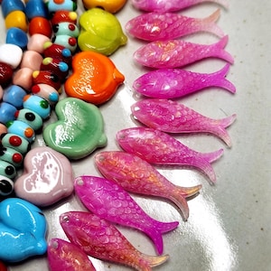 May include: A collection of colorful glass beads and charms in various shapes, including hearts, fish, and abstract forms. The beads are in shades of pink, orange, yellow, blue, and green. The fish charms have a translucent, iridescent quality.
