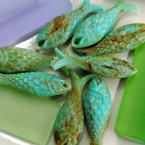 May include: A collection of turquoise and brown fish-shaped glass beads. The beads have detailed scale and fin designs, with a hole for stringing. Various green and purple glass pieces are scattered around the fish.