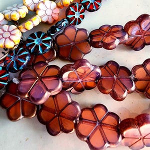 May include: Close-up of several strands of colorful beads. The beads feature various floral designs in shades of pink, brown, red, blue, and yellow. The beads are strung together, showcasing their intricate details and vibrant colors.
