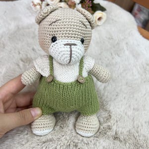Op de afbeelding: Handgemaakte gehaakte teddybeer met groene tuinbroek met houten knoopdetails. De beer heeft een beige kop, witte snuit en wit shirt. De ogen van de beer zijn zwart en de tuinbroekriemen zijn groen.