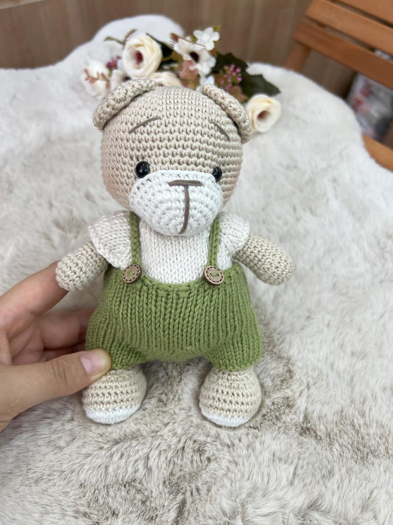 Op de afbeelding: Handgemaakte gehaakte teddybeer met groene tuinbroek met houten knoopdetails. De beer heeft een beige kop, witte snuit en wit shirt. De ogen van de beer zijn zwart en de tuinbroekriemen zijn groen.