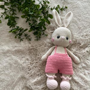 May include: A crocheted bunny doll with a light beige body, long ears, and pink cheeks. The bunny is dressed in pink dungarees and matching booties. The doll is on a soft, textured, light brown surface.
