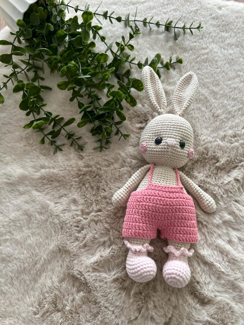 May include: A crocheted bunny doll with a light beige body, long ears, and pink cheeks. The bunny is dressed in pink dungarees and matching booties. The doll is on a soft, textured, light brown surface.