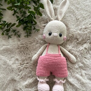 May include: A handmade crochet bunny doll with a cream-coloured body, long ears, and pink cheeks. The bunny wears pink shorts with braces and matching ruffled socks. The doll is approximately 25 cm tall.