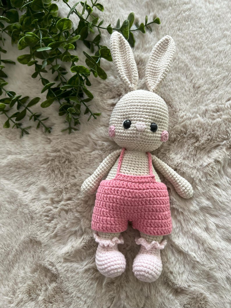 May include: A handmade crochet bunny doll with a cream-coloured body, long ears, and pink cheeks. The bunny wears pink shorts with braces and matching ruffled socks. The doll is approximately 25 cm tall.