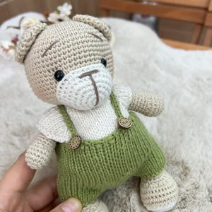 Op de afbeelding: Een handgemaakte gehaakte teddybeer met een lichtbruine kop en ledematen, een wit gezicht en shirt en een groene tuinbroek. De beer heeft zwarte ogen en een bruin gestikte neus en mond. De tuinbroek heeft houten knoopdetails.