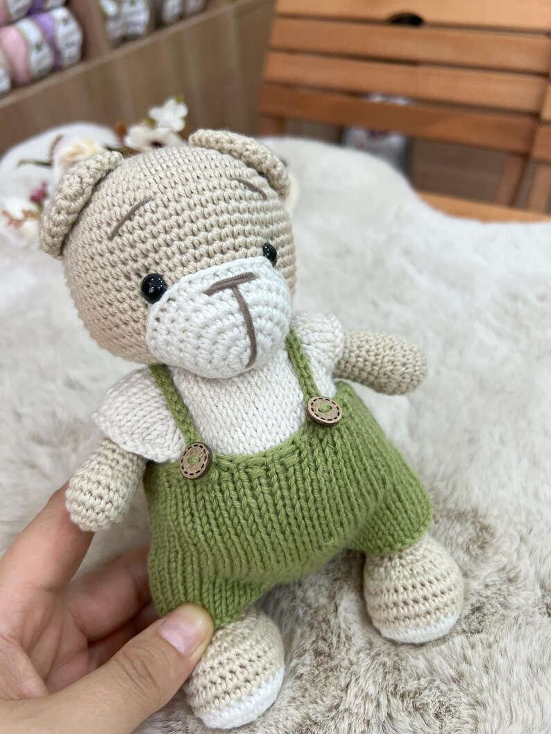 Op de afbeelding: Een handgemaakte gehaakte teddybeer met een lichtbruine kop en ledematen, een wit gezicht en shirt en een groene tuinbroek. De beer heeft zwarte ogen en een bruin gestikte neus en mond. De tuinbroek heeft houten knoopdetails.