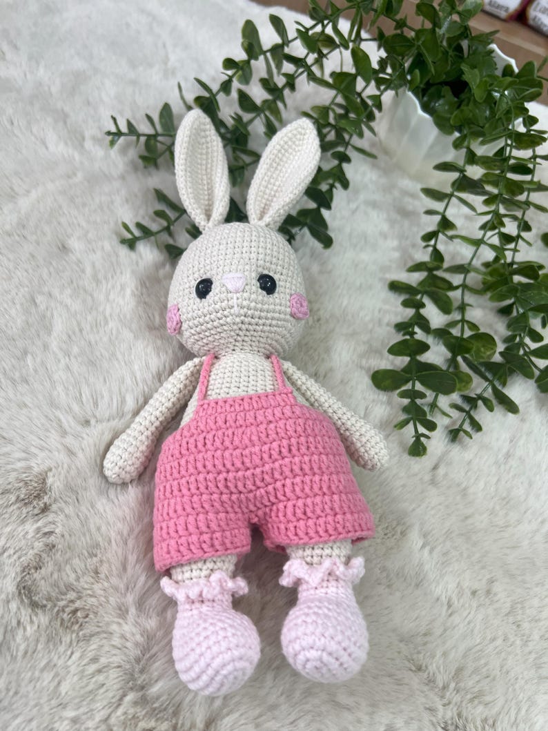 May include: A handmade crochet bunny doll with a cream body, long ears, and pink cheeks. The bunny wears pink overalls and matching ruffled booties. The doll is a soft toy, suitable for children.