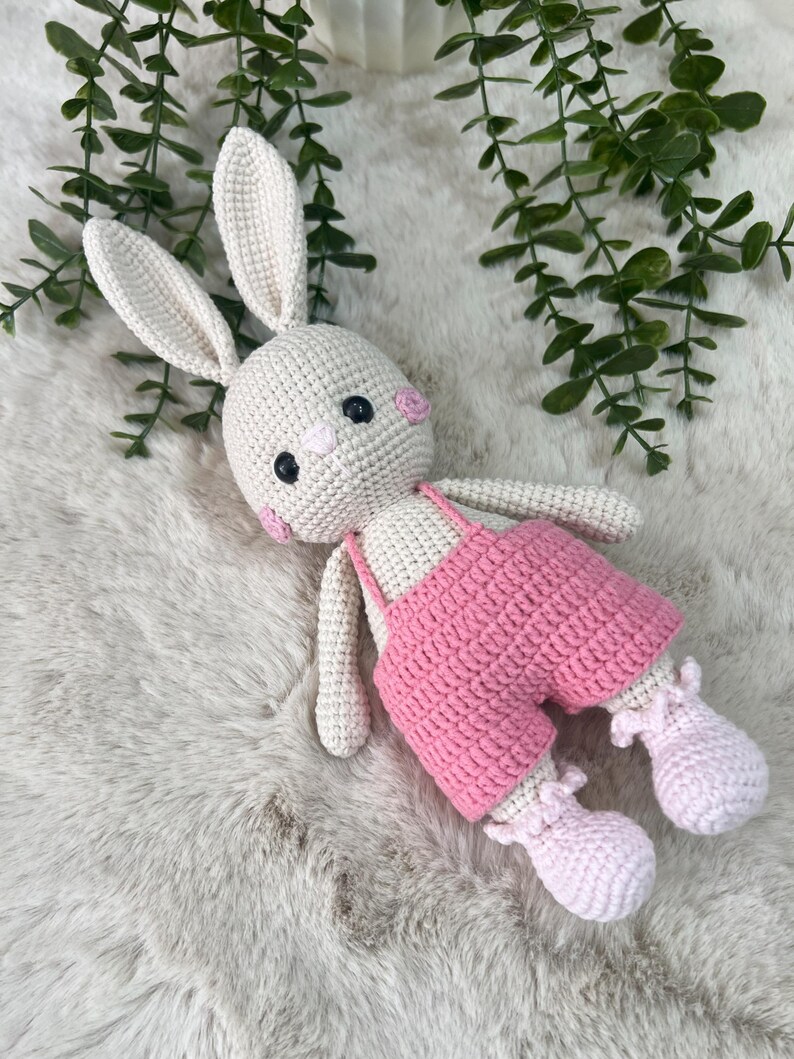 May include: A handmade crochet bunny doll with a cream body, long ears, and pink cheeks. The bunny wears pink dungarees and matching booties. The doll is on a soft, white surface, with green foliage in the background.