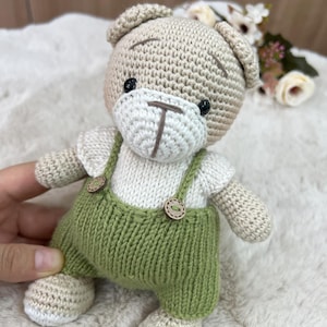 Op de afbeelding: Een handgemaakte gehaakte teddybeer met groene tuinbroek met houten knopen. De beer heeft een beige hoofd, armen en benen, een witte gezicht en shirt en zwarte ogen. De beer is een zacht speelgoed.