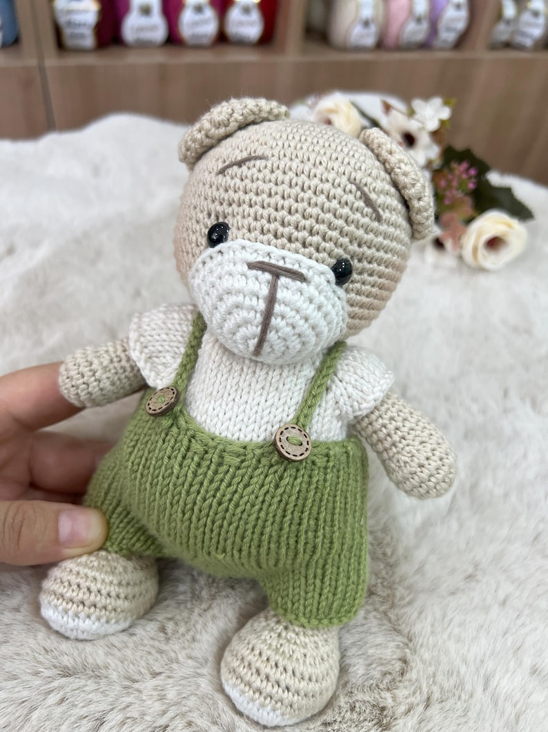 Op de afbeelding: Een handgemaakte gehaakte teddybeer met groene tuinbroek met houten knopen. De beer heeft een beige hoofd, armen en benen, een witte gezicht en shirt en zwarte ogen. De beer is een zacht speelgoed.