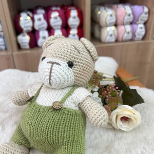 Op de afbeelding: Een handgemaakte gehaakte teddybeer met een beige lichaam, een wit shirt en een groene overall. De beer heeft zwarte ogen en houten knoopdetails. De beer zit op een witte, pluizige ondergrond.