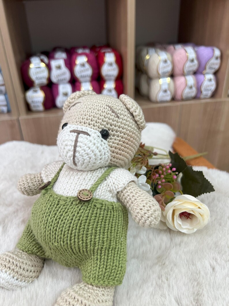Op de afbeelding: Een handgemaakte gehaakte teddybeer met een beige lichaam, een wit shirt en een groene overall. De beer heeft zwarte ogen en houten knoopdetails. De beer zit op een witte, pluizige ondergrond.