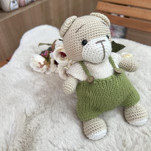 Op de afbeelding: Een handgemaakte gehaakte teddybeer met groene tuinbroek met houten knopen. De beer heeft een beige lichaam, een wit gezicht en zwarte ogen. Hij zit op een zacht wit oppervlak met bloemen.