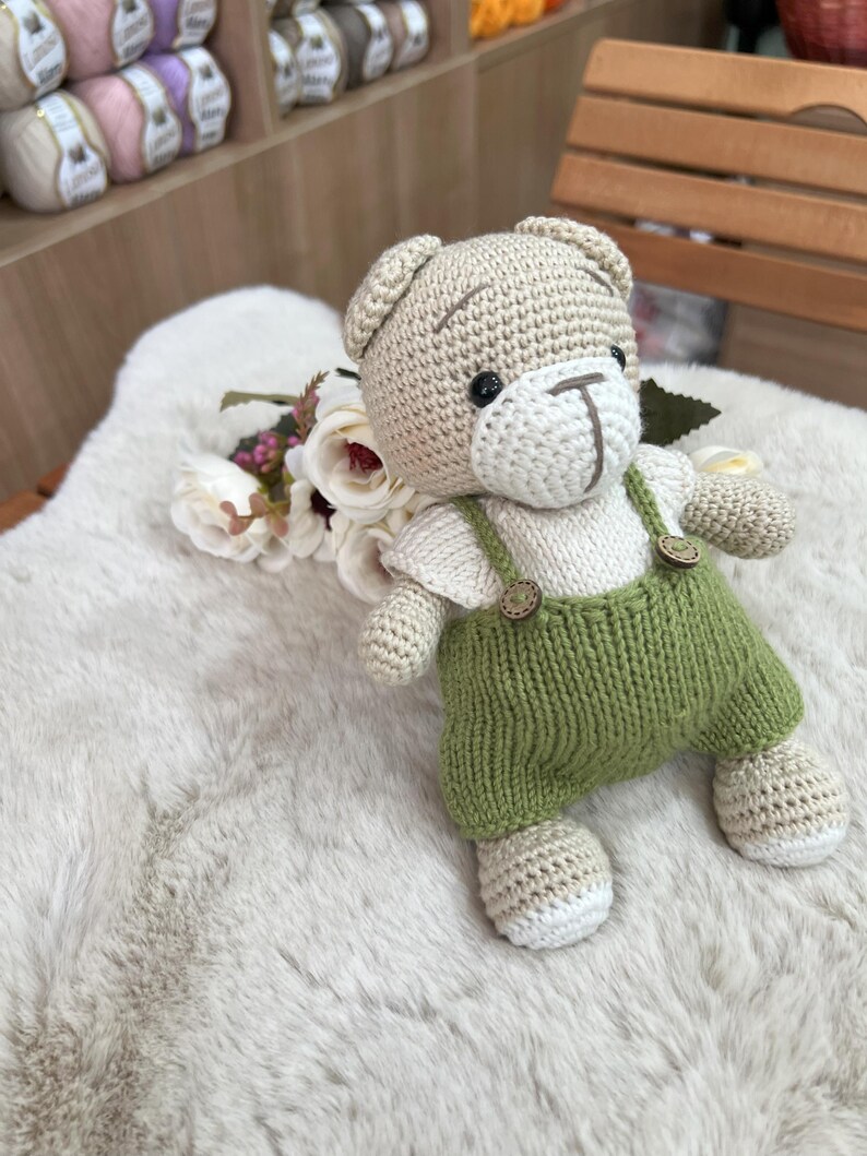 Op de afbeelding: Een handgemaakte gehaakte teddybeer met groene tuinbroek met houten knopen. De beer heeft een beige lichaam, een wit gezicht en zwarte ogen. Hij zit op een zacht wit oppervlak met bloemen.