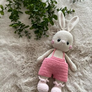 May include: A handmade crochet bunny toy, featuring a cream-coloured body, pink cheeks, and black eyes. It's dressed in pink dungarees and matching ruffled booties. The bunny rests on a fluffy, light-toned surface, with green foliage visible.