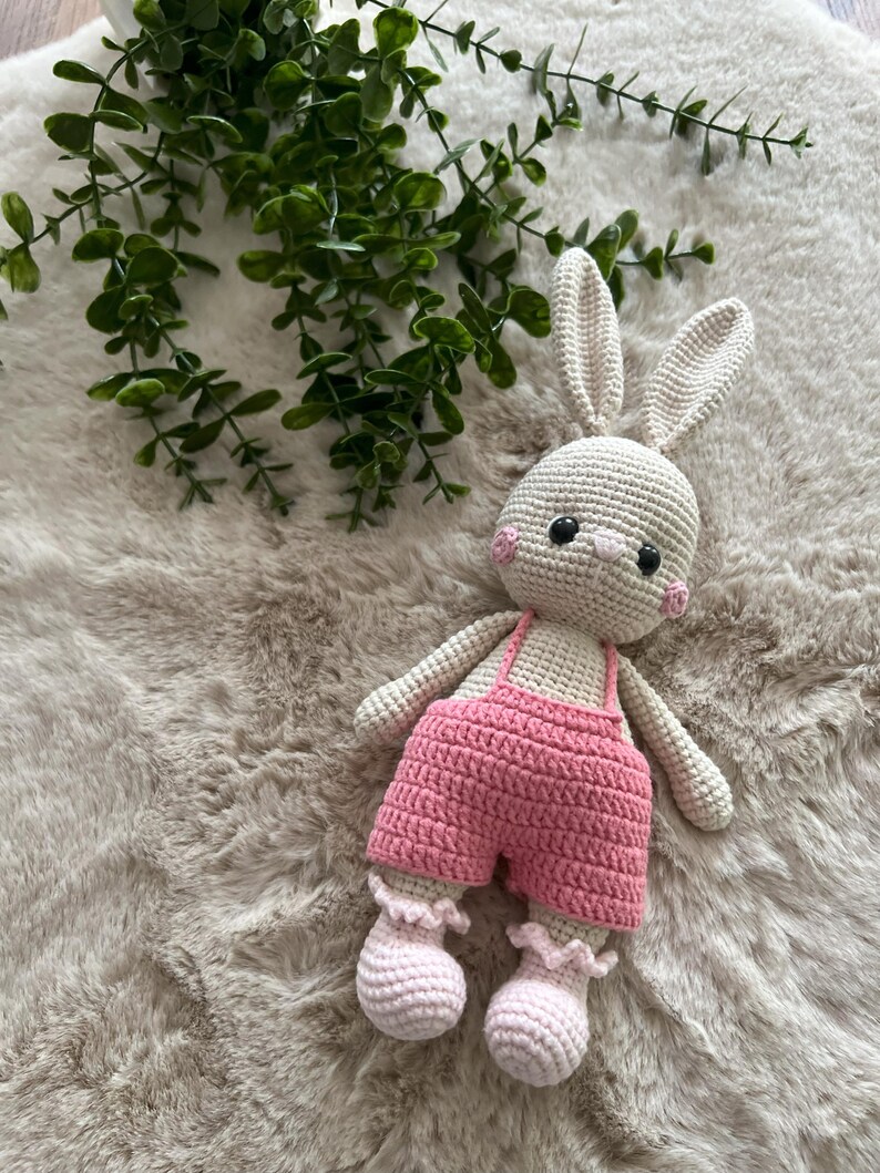 May include: A handmade crochet bunny toy, featuring a cream-coloured body, pink cheeks, and black eyes. It's dressed in pink dungarees and matching ruffled booties. The bunny rests on a fluffy, light-toned surface, with green foliage visible.