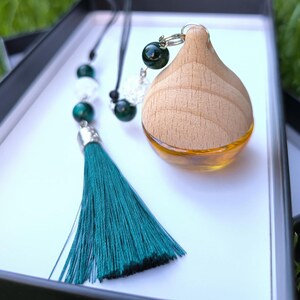 Könnte beinhalten: Ein ätherisches Öl-Diffusor aus Holz mit einer Glasbasis, die mit bernsteinfarbener Flüssigkeit gefüllt ist. Er hat eine türkisfarbene Quaste und Perlenakzente. Der Diffusor wird in einer weißen Box präsentiert.