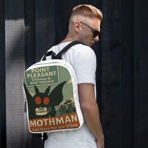 Może przedstawiać: Biały plecak z motywem Mothmana i napisem "POINT PLEASANT Welcome to WEST VIRGINIA" oraz "MOTHMAN Just Here for the Vibes". Plecak ma czarne paski i czarny zamek błyskawiczny.