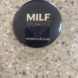 May include: A black button featuring the word "MILF" in white capital letters. Below is a mothman graphic with red eyes and wings. The text "Mothman I'd like to find" is printed below the graphic.