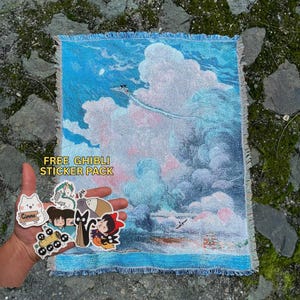 Puede incluir: Un tapiz azul y blanco con un diseño de paisaje de nubes, que presenta una escena con una figura voladora. El tapiz tiene un borde con flecos. Se incluye un paquete de pegatinas Ghibli gratis con varias pegatinas de personajes.