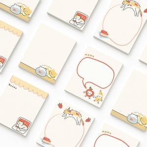 May include: A collection of cream-colored memo pads with various illustrations. Some pads feature a sleeping cat, a speech bubble, or the text "Let's play" and "Priorities." The pads have a scalloped or straight edge design.