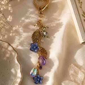 May include: A gold-coloured keychain with a heart-shaped clasp. The charm features blue flowers, gold leaves, and iridescent and purple teardrop-shaped pendants. A small gold crescent moon and a pearl are also attached.