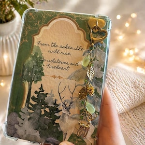 May include: A clear phone case with a forest scene and the quote "From the ashes she will rise." The case features a deer, trees, and a gold charm with leaf and deer pendants. The case is approximately 15 cm tall.