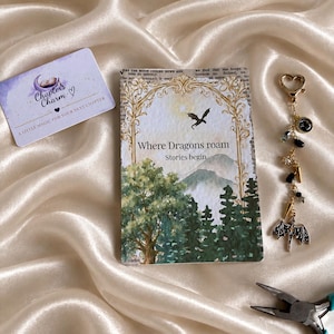 May include: A rectangular card featuring a forest landscape, a dragon silhouette, and the text Where Dragons roam Stories begin, accompanied by a heart-shaped clasp keychain with black and gold beads and a dragon charm, plus a small branded card.