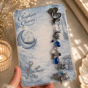 May include: A clear protective phone case featuring a light blue background with a crescent moon, an open book, and the text Chapters Charm. A silver-toned metal charm dangles from the top, adorned with a heart, a moon, blue beads, a star, and a bat.