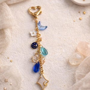 May include: A celestial-themed keyring with a gold heart-shaped clasp. The charm features a blue crescent moon, white flower, a navy blue charm with a moon and stars, and various blue and clear crystal charms. A gold star-shaped charm completes the design.
