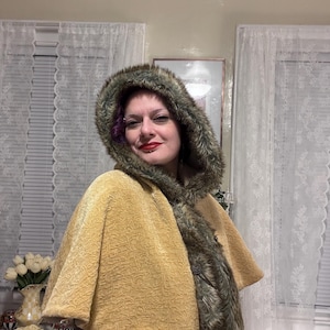 May include: A mustard yellow cape with a textured pattern and a faux fur-lined hood. The hood and trim feature shades of brown and green. The cape has a button closure and a classic design.