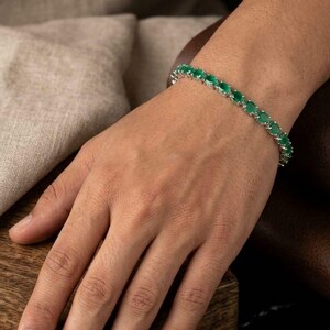 May include: A silver bracelet with a row of oval-shaped, bright green gemstones. The bracelet is worn on a wrist, highlighting the design and colour of the jewellery. The gemstones are set close together, forming a continuous line of green.