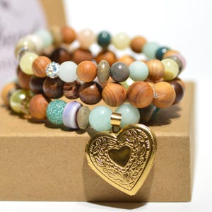 May include: A multi-strand bracelet featuring a mix of round wooden, smooth stone, and textured beads in shades of brown, green, and cream. A gold-toned heart-shaped locket with an embossed floral pattern hangs from the bracelet as a central charm.