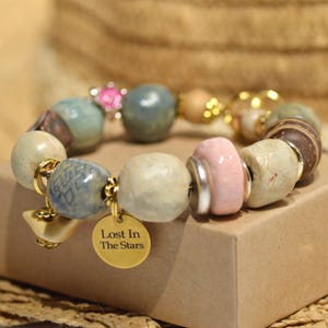 Può includere: Un braccialetto di perline con una varietà di perle in ceramica e metallo nei toni del blu, rosa, beige e marrone. Il braccialetto include un ciondolo rotondo color oro inciso con il testo Lost In The Stars e un piccolo ciondolo a forma di uccello.