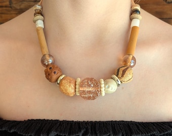 May include: A necklace featuring a central faceted translucent peach bead flanked by textured tan, brown, and cream-colored beads. The design includes smooth wooden cylinders, metallic silver spacers, and small round beads in various earthy tones.