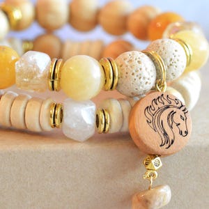 May include: A multi-strand bracelet featuring a mix of light wood beads, smooth yellow stones, textured white lava beads, and clear faceted crystals. Gold-toned metal spacers separate the beads, and a round wooden charm with a horse head engraving.