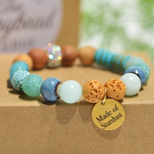 May include: A beaded bracelet with a variety of colorful beads, including turquoise, brown, and white. The bracelet has a gold charm that reads "Made of Stardust". The beads are arranged in a circular pattern.