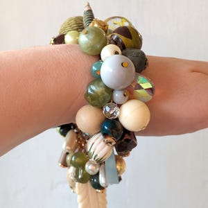 Puede incluir: Una pulsera gruesa con una variedad de cuentas en tonos verdes, crema, marrón y dorado. La pulsera presenta cuentas de madera, vidrio e iridiscentes, junto con un dije de hoja tallada. Las cuentas están ensartadas, creando un accesorio único y texturizado.