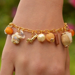 Puede incluir: Pulsera dorada con dijes decorativos variados. Los dijes incluyen cuentas naranjas, una estrella de mar, cristales transparentes, un pequeño objeto dorado, una perla y elementos de madera y piedra. La pulsera se lleva en la muñeca.