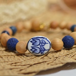 May include: A beaded bracelet featuring wooden beads, blue lava beads, and a central white ceramic bead with a blue floral design. The bracelet is set against a woven straw background.