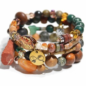 Könnte beinhalten: Ein mehrreihiges Wickelarmband mit einer Auswahl an Perlen in Braun-, Grün-, Orange- und Schwarztönen. Das Armband enthält Holz-, Stein- und Glasperlen sowie einen runden goldfarbenen Anhänger, der mit einem Unendlichkeitssymbol graviert ist.